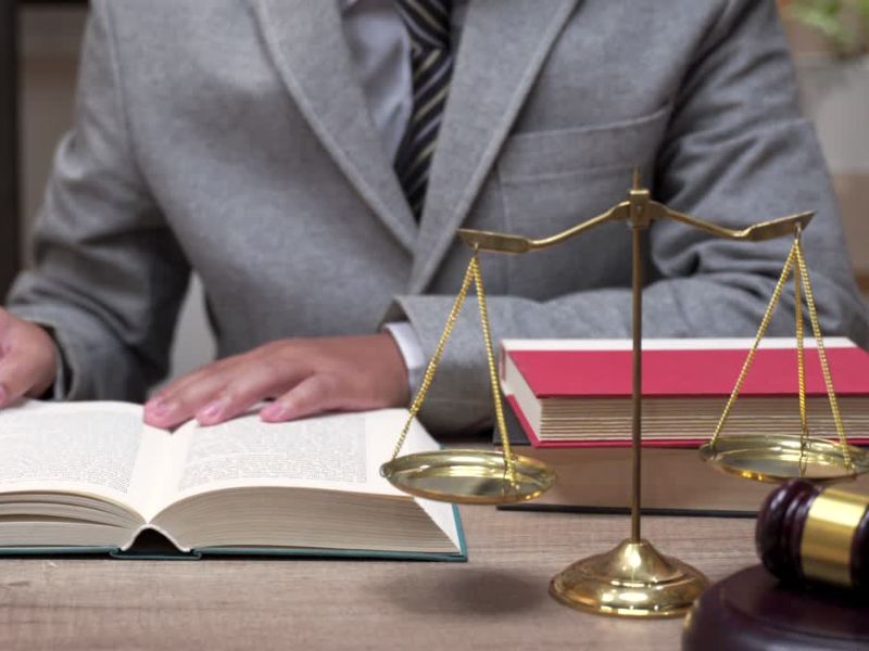 Osoba u poslovnom odelu sedi za stolom i čita pravnu knjigu. Na stolu se nalaze vaga pravde, čekić sudije i još nekoliko knjiga, što simbolizuje pravni sistem i pravnu praksu.
