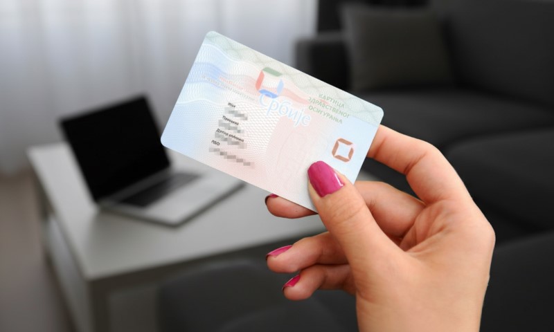 Osoba drži zdravstvenu knjižicu u ruci, laptop u pozadini na sredini dnevne sobe