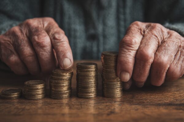 Starije ruke slažu male gomile kovanica na drvenom stolu, simbolizujući penziju i finansijsku sigurnost u starosti