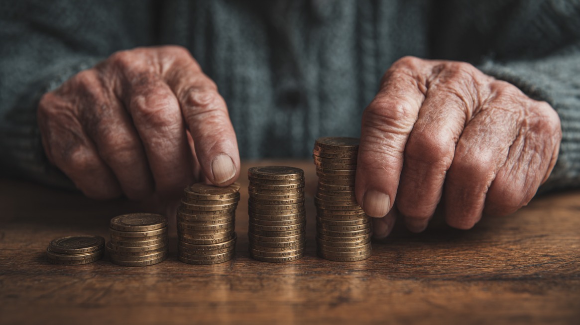Starije ruke slažu male gomile kovanica na drvenom stolu, simbolizujući penziju i finansijsku sigurnost u starosti