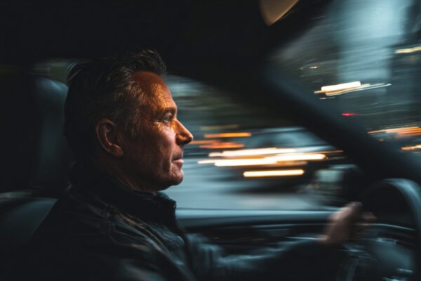 Muškarac srednjih godina sedi za volanom automobila i vozi noću, sa zamućenim svetlima grada u pozadini
