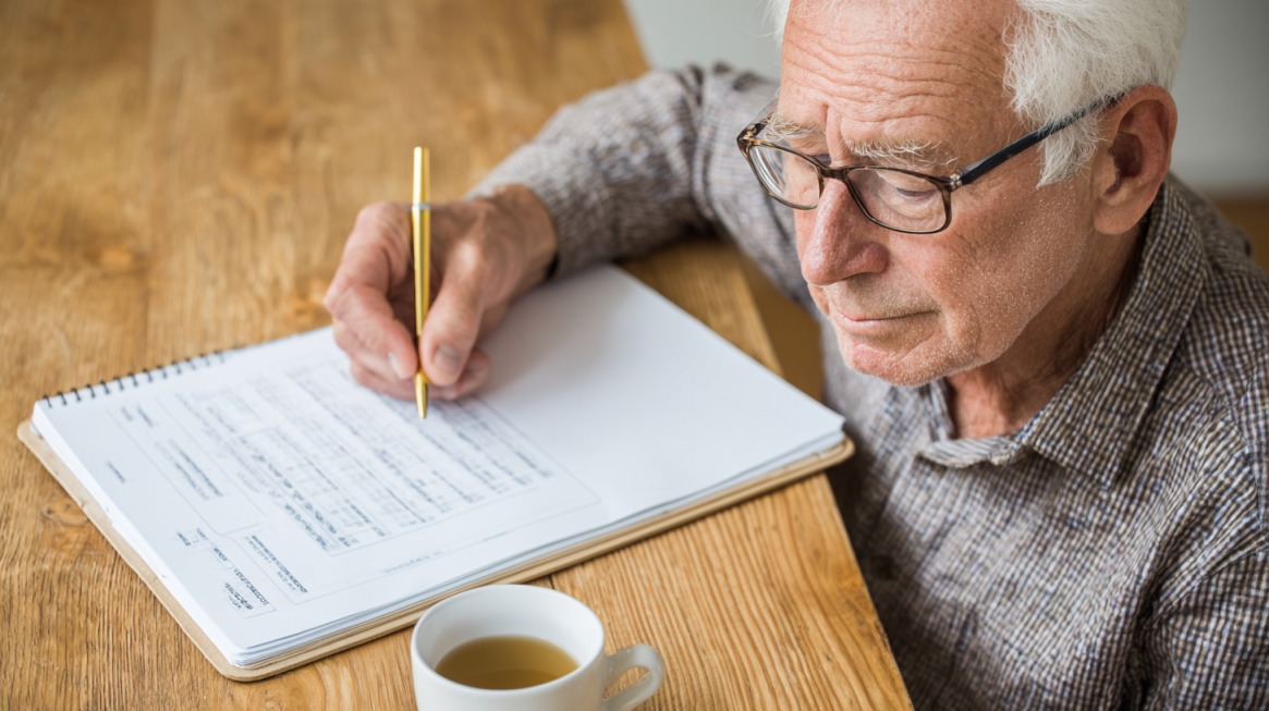 Stariji muškarac sa naočarima sedi za stolom, proučava dokumente i zapisuje podatke u svesku, uz šolju čaja.