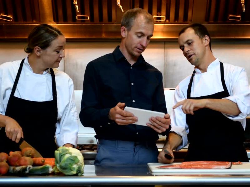 dva kuvara i vlasnik resotorana prave meni u kuhinji dok radnici seku ribu i povrće