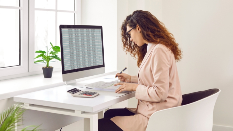 Knjigovođa u kancelariji radi za računarom, pregleda tabelu sa podacima i beleži finansijske informacije na papiru