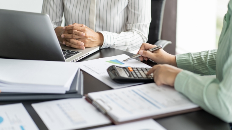 Dve osobe za stolom pregledaju finansijske izveštaje, koriste kalkulator i laptop tokom poslovnog sastanka