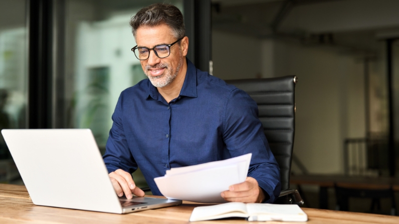 Muškarac u kancelariji pregleda dokumenta i radi na laptopu za radnim stolom
