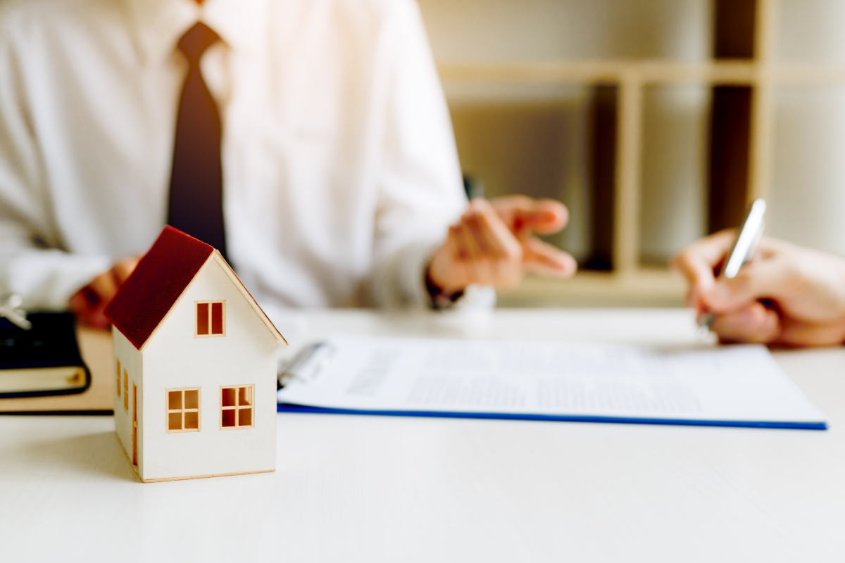 osoba popunjava papire za promenu vlasništva stana, pored njega maketa drvene kućice i druge osobe koja drži predaje ugovor 