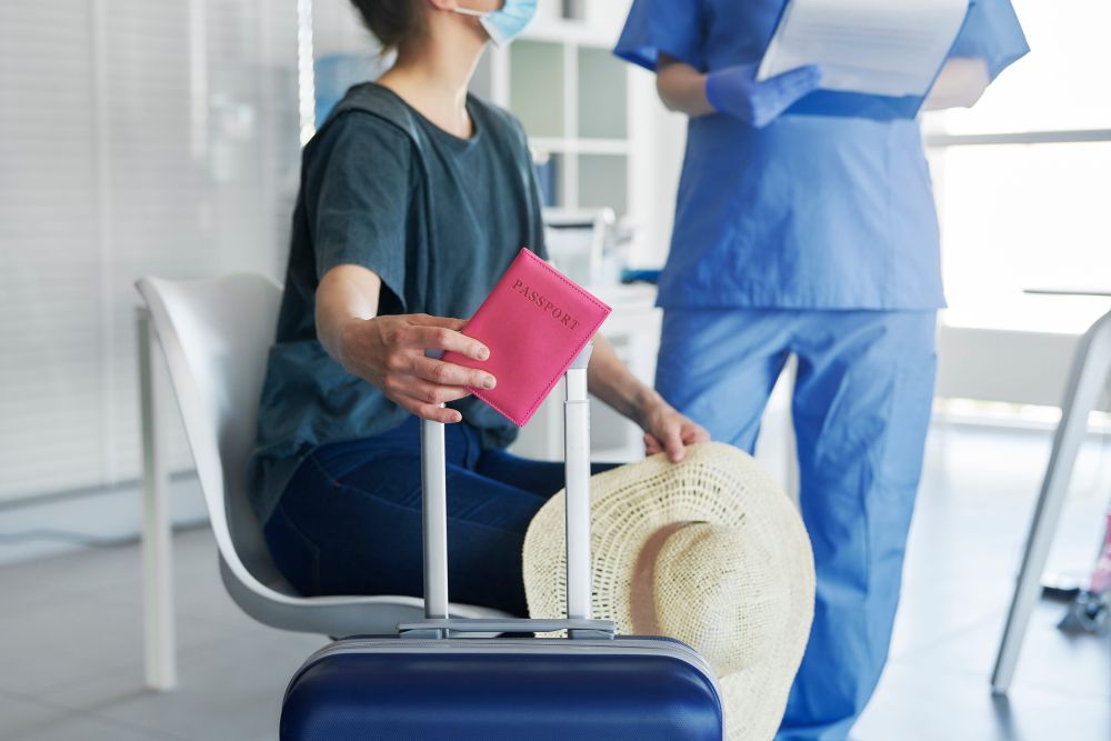 Putnica sa pasošem i koferom razgovara sa medicinskim osobljem u zdravstvenoj ustanovi.