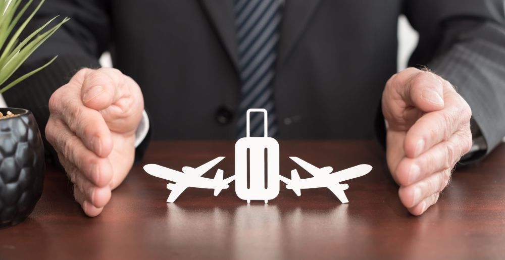 Osiguravajući agent rukama štiti simbole kofera i aviona koji predstavljaju putno osiguranje.